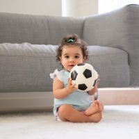 Happy black curly haired baby girl in pale blue clothes sitting on floor at home, looking away, playing soccer ball. Front view. Kid at home and childhood concept