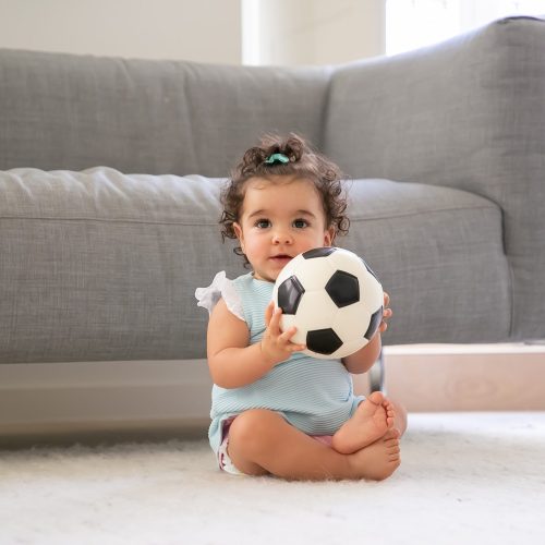 Happy black curly haired baby girl in pale blue clothes sitting on floor at home, looking away, playing soccer ball. Front view. Kid at home and childhood concept